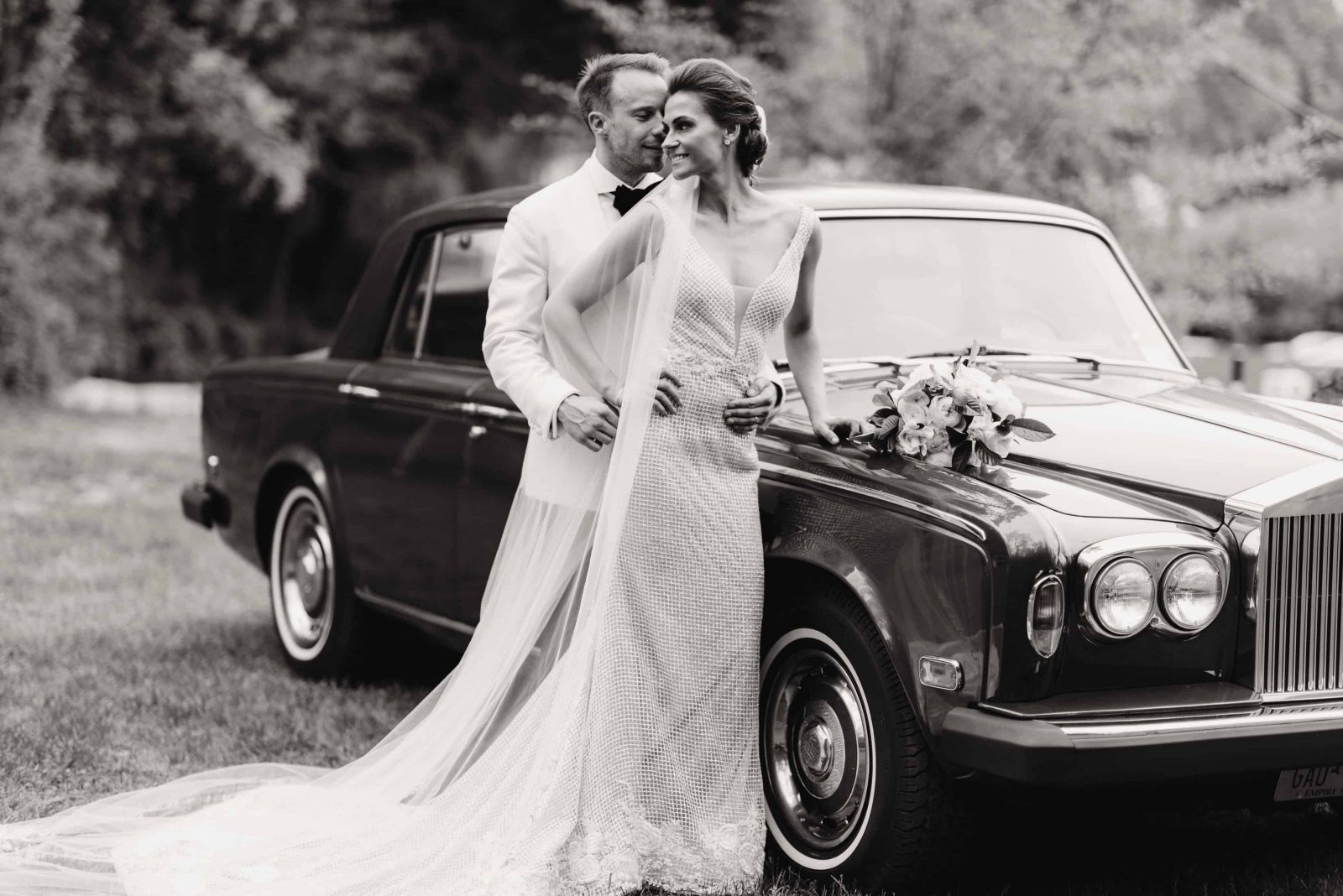 Black and white photo of newlyweds standing next to a classic car